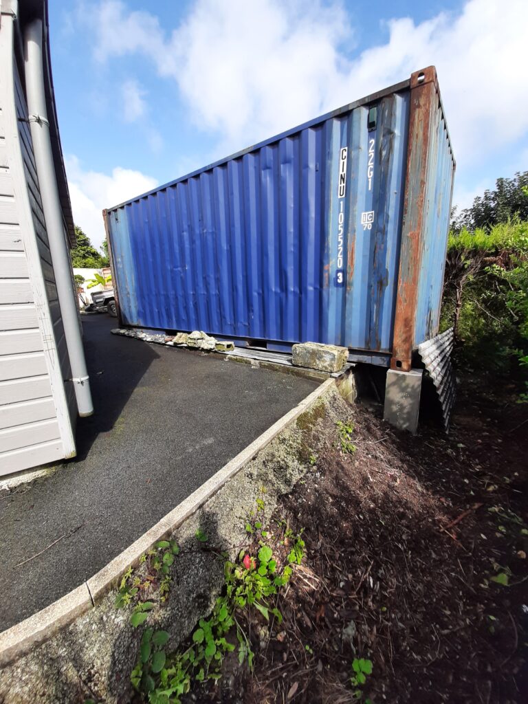 Un container maritime bleu de 20 pieds installé près d'une maison, légèrement surélevé avec des blocs pour stabilisation, entouré d'un sol aménagé.