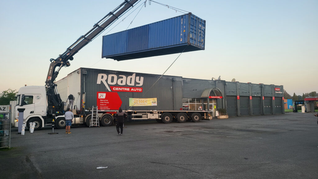 Container maritime bleu de 40 pieds en cours de levage par une grue devant la façade du centre auto ROADY de Montpon-Ménestérol, solution de stockage In'Box Container.