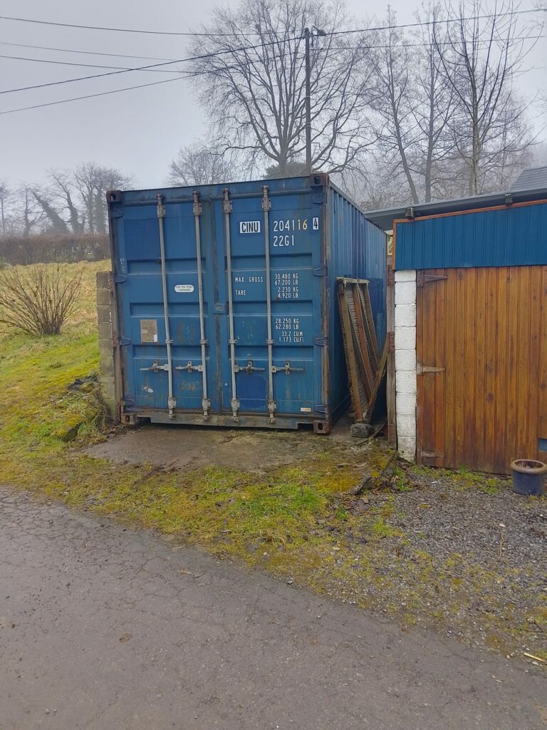 Container garage bleu installé dans espace restreint à Manhay avec maison en arrière-plan